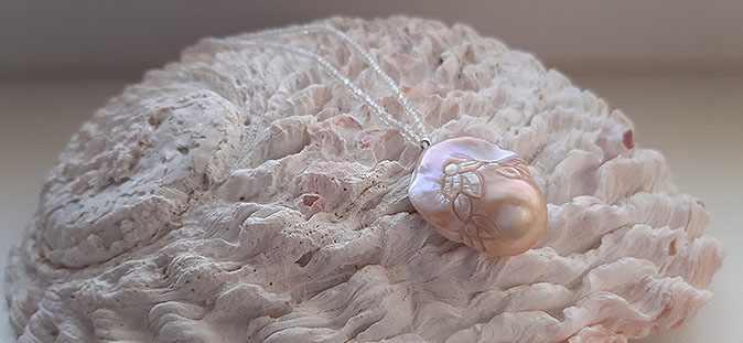 Eine mit Kalk bewachsene weiß-rosa Muschel auf der eine Kette aus klarem, facettiertem Zirkon liegt. In der Mitte der Kette befindet sich eine große, rosa-apricot-schimmernde Edisonperle, die kunstvoll mit Blütenblättern graviert wurde.