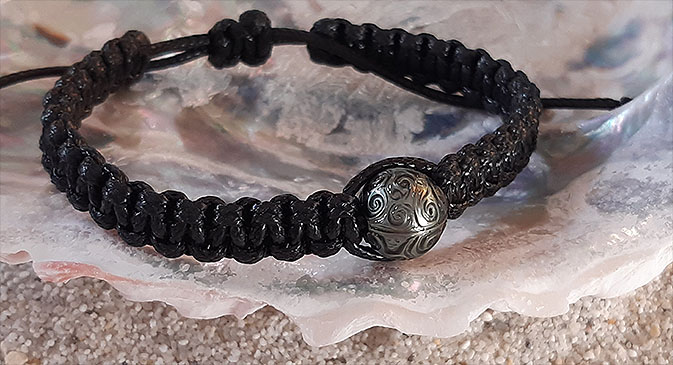 Eine Muschel, die im Sand liegt. In ihrem Innern liegt ein aus Makramee geflochtenes schwarzes Armband in das eine gravierte olivgrüne Marutea-Perle eingebunden ist. 